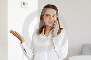 Indoor shot of young adult confused woman wearing white casual style shirt, posing in light room at home, talking via phone with