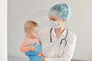 Indoor shot of young adult female pediatrist wearing medical uniform, holding infant baby girl, examining child`s health in clini