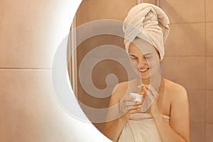 Indoor shot of young adult female taking care of her skin, posing in bathroom in front of mirror with cosmetic cream in hands,
