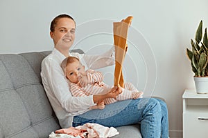 Indoor shot of young adult mother sitting on sofa with her infant daughter, showing new attire for her charming kid, looking at