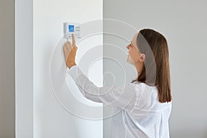 Indoor shot of young adult woman regulating heating temperature with a modern wireless thermostat installed on the white wall at