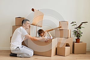 Indoor shot of young brunette man wearing casual attire sitting on floor and playing with his cute dark haired daughter, little