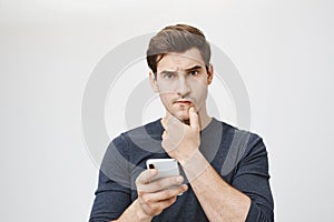 Indoor shot of young caucasian student looking worried and perplexed while holding smartphone and touching ching