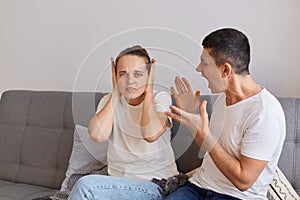 Indoor shot of young couple having conflict, angry fury man sitting on sofa and yelling on his wife with raised arms, having