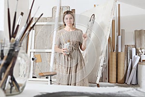 Indoor shot of young female artist looking at camera standing in front of the canvas holding painting tools in bright white studio