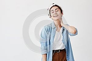 Indoor shot of young female with dark and wavy hair in bun wears denim shirt and brown trousers, with hand on neck, uses