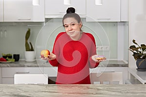 Indoor shot of young pregnant woman with apple and tasty cake in her hands, decides what to eat, attractive female posing in house