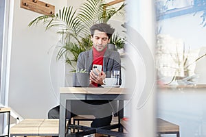 Indoor shot of young pretty dark haired bearded guy with trendy haircut sitting over modern cafe interior and looking on his