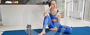 Indoor shot of young sportswoman holding smartphone, sits with fitness water bottle, uses sports app to workout, do yoga