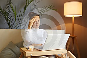 Indoor shot of young woman doing research work for her business while sitting on sofa, sad tired girl browsing through the