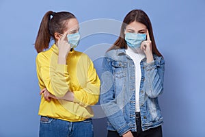 Indoor shots of two thoughtful females having antibacterial masks on face, touching temples with fingers, having pensive facial