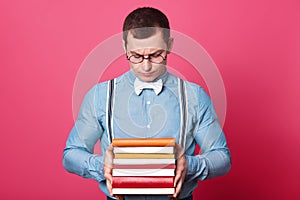 Indoor studio shot of dark haired confused model posing isolated over bright pink background, holding books in his strong hands,