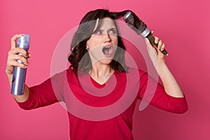 Indoor studio shot of screaming annoyed brunette, styling her hair, comb tangling hair, holding hairbrush and spray in both hands