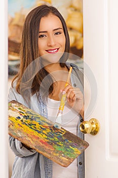 Indoor view of beautiful young woman painter with color palette and paint brush in hand, in a blurred background