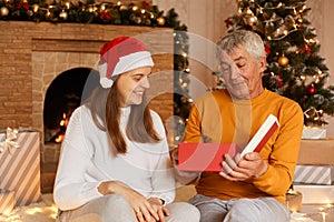 Indor shot of happy family young woman and her senior father sitting in living room with fireplace and Christmas tree, opening new