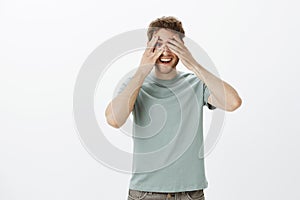 Indor shot of playful happy mature male model in t-shirt, covering eyes with palms and peeking through fingers, smiling