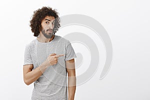 Indor shot of unaware doubtful attractive hispanic guy with beard and afro hairstyle, pointing right with forefinger and