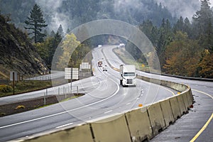 Industrial big rig semi truck with semi trailer transporting cargo driving on the winding highway road with fog and forest