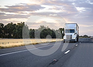 Industrial bonnet American big rig semi truck with covered dry van semi trailer driving on the evening twilight highway road with