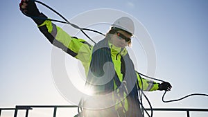 Industrial climber in a white helmet coils a rope on a roof against the backdrop of the sun