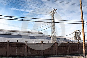Industrial complex behind a tall stucco wall topped with barbed wire against a blue sky with clouds, urban industrial landscape