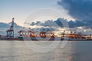 Industrial Container Cargo freight ship with working crane bridge in shipyard at dusk for Logistics Import Export background.