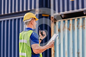 Industrial engineer in hardhat containers box background, Dock worker man talks on two-way radio with holding clipboard checklist
