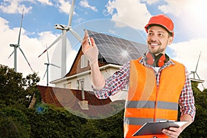 Industrial engineer in uniform and view of wind energy turbines near house with installed solar panels on roof