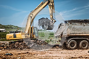 Industrial excavator using scoop and moving earth and trash at garbage dumping site.