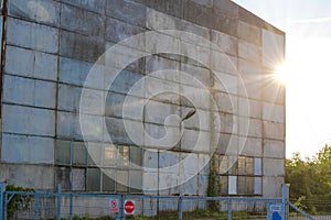 An industrial hall with the setting sun peeking out from behind its edge. Signs on the gate: \