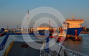 Industrial harbor with moored large ships with masts, barges with cargo, boats in red light of sunset. General view from ferry