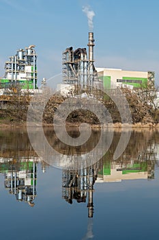 Industrial landscape. A large factory with pipes is reflected in the clear river water against the blue sky. Toxic smoke is coming