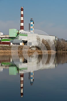 Industrial landscape. A large factory with pipes is reflected in the clear river water against the blue sky. Toxic smoke is coming