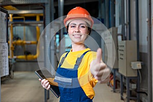 Industrial production. A female engineer in uniform and a protective helmet, holding a tablet in her hands, gives a thumbs up. The