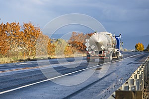 Industrial standard blue big rig semi truck transporting liquid cargo in tank semi trailer running on the winding autumn highway