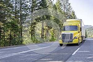 Industrial standard yellow big rig bonnet semi truck transporting goods in dry van semi trailer driving on the interstate highway
