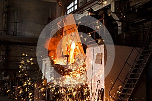 Industrial worker oversees molten metal pouring from furnace at steel mill. Foundry specialist in protective gear