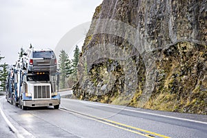 Industry standard big rig semi truck car hauler transporting cars running on the winding mountain road with rock wall on the side