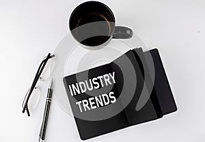 INDUSTRY TRENDS written text in small black notebook with coffee , pen and glasess on white background. Black-white style