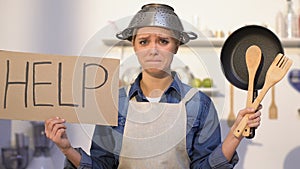 Inexperienced lady with colander on head holding kitchen utilities and help sign