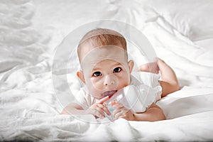 Infant baby with big brown eyes lying on the white bed and licks special bottle of water with nipple. Tries to gnaw her. teething