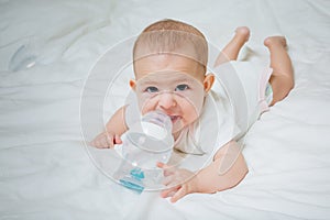 Infant baby with big brown eyes lying on the white bed with special bottle of water with nipple. Tries to gnaw her. teething