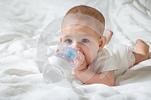 Infant baby with big brown eyes lying on the white bed with special bottle of water with nipple. Tries to gnaw her. teething