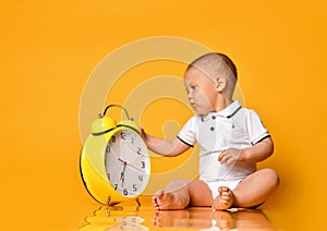 Infant baby boy toddler is occupied with a big yellow alarm clock playing looking exploring on yellow background
