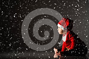 Infant baby boy toddler in red christmas cap and new year costume is sitting on ice under the snow staring at light ray