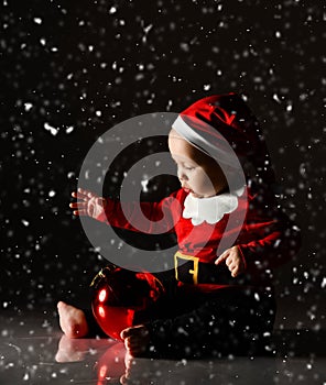 Infant baby boy toddler is sitting in santa claus costume, cap under the snow playing with big red christmas tree ball
