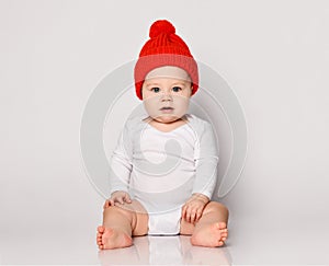 Infant baby boy toddler in white bodysuit and red warm hat with pompon is sitting looking directly at camera on white