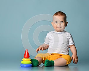 Infant baby boy toddler in yellow pants and white t-shirt is playing with a children multi-colored developing pyramid