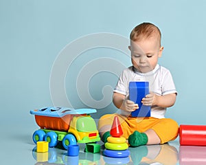 Infant baby boy toddler in yellow pants and white t-shirt is sitting on the floor surrounded by colorful toys