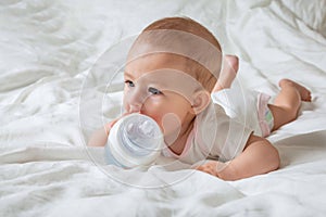 Infant baby girl lying on the white bed with special bottle of water with nipple. Tries to gnaw her. Aching teeth